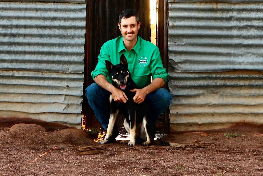 A man in a long-sleeved shirt sits with a dog on a farm.