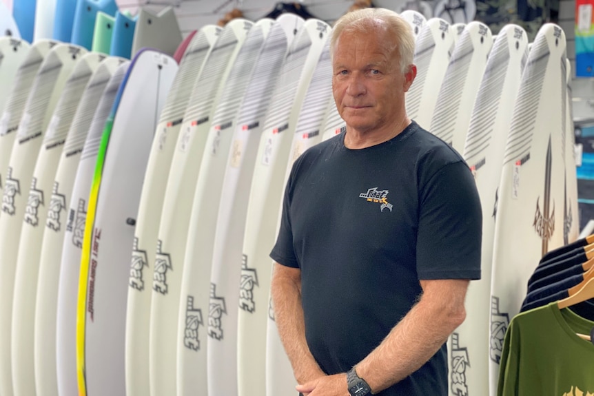 Man with short hair stands in front of a row of surf boards, looking stern and not smiling
