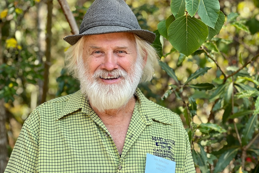 A bearded man in a hat smiles in front of young trees.