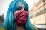 A woman with blue hair and a maroon face mask looks straight at the camera