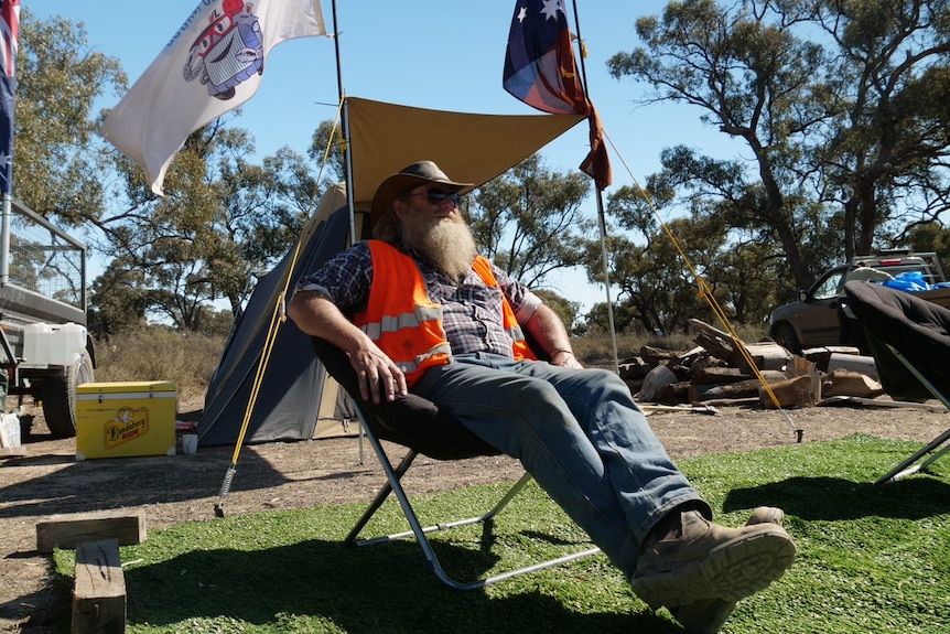 A bearded man in a leather cowboy hat sits in a deckchair surrounded by flags.