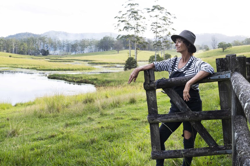 A smiling older woman in a hat and overalls leans on a gate and looks out at a country property.