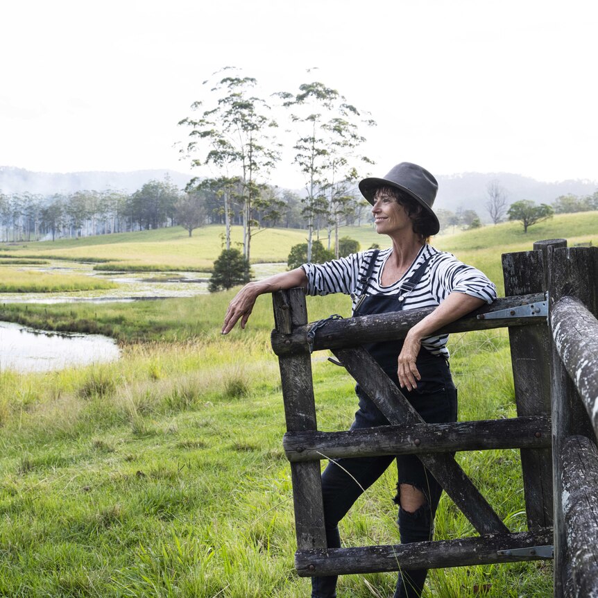 A woman in overalls and a hat is leaning on a wooden gate, looking out over a pond and paddock