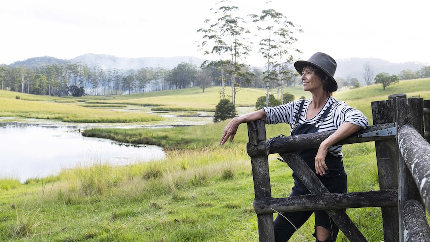 A woman in overalls and a hat is leaning on a wooden gate, looking out over a pond and paddock