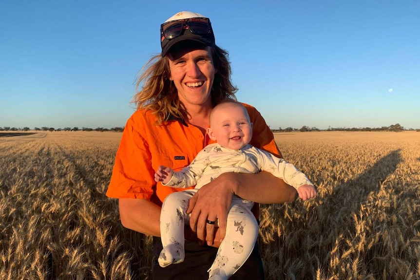 A man wears an orange hi-vis shirt and a cap with sunglasses on it as he holds a smiling baby while standing in a field.