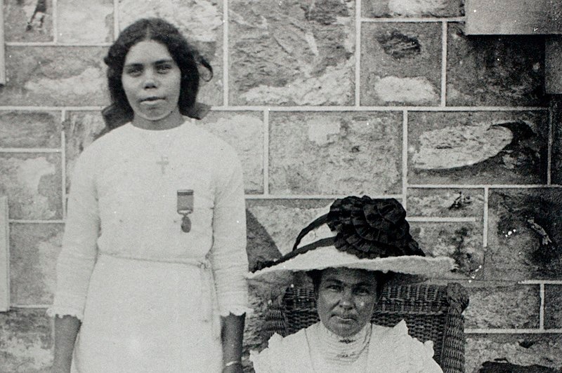 Cissy McLeod standing with bravery medal pinned to dress, her employer she saved from drowning sits next to her in chair.