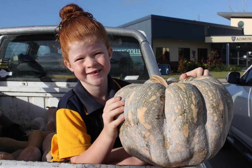 A young girl sitting in a ute tray full of pumpkins holds a large pumpkin up
