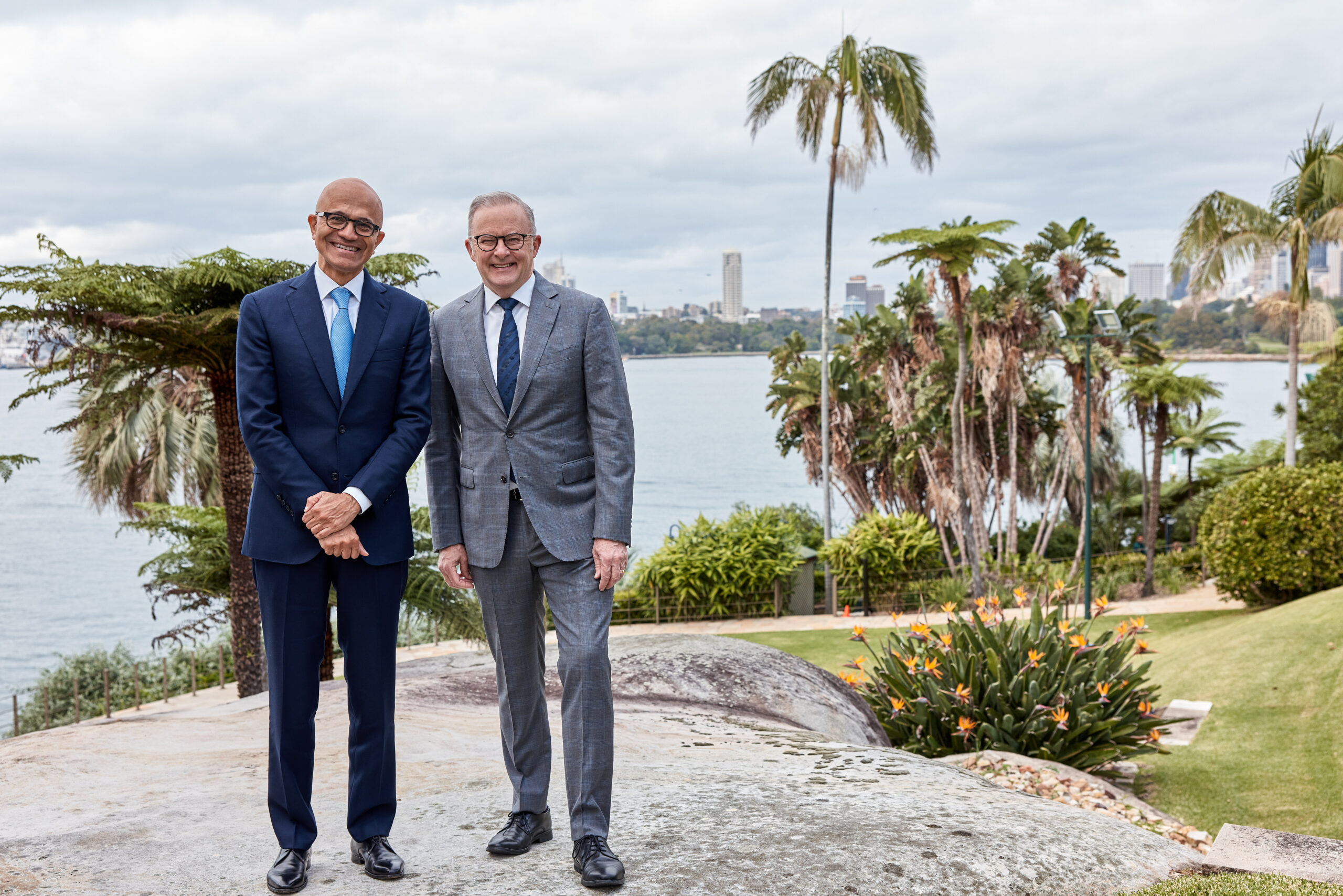 Two men wearing suits stand on a rock in front of the Sydney city skyline on a sunny day.