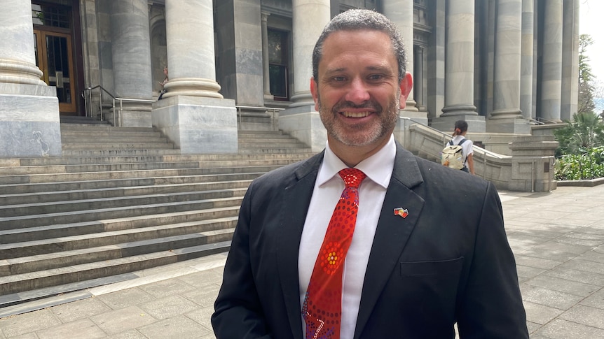 An aboriginal man in a suit stands outside a grey parliament building