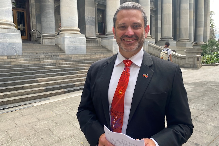 An aboriginal man in a suit stands outside a grey parliament building