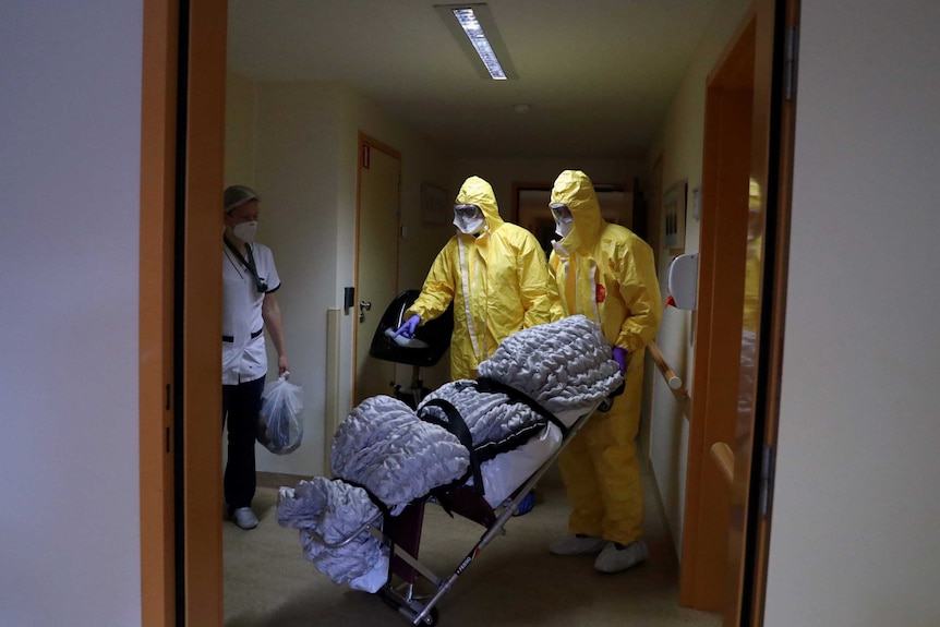 Two health workers in PPE wheeling out a body wrapped in a blanket