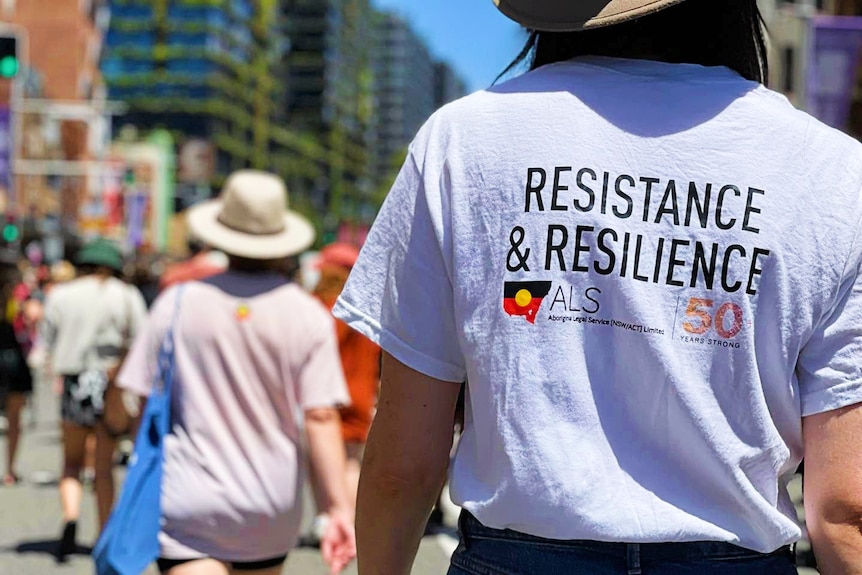 the  back of a person at a rally with a t shirt that reads resistance and resilience with an aboriginal flag