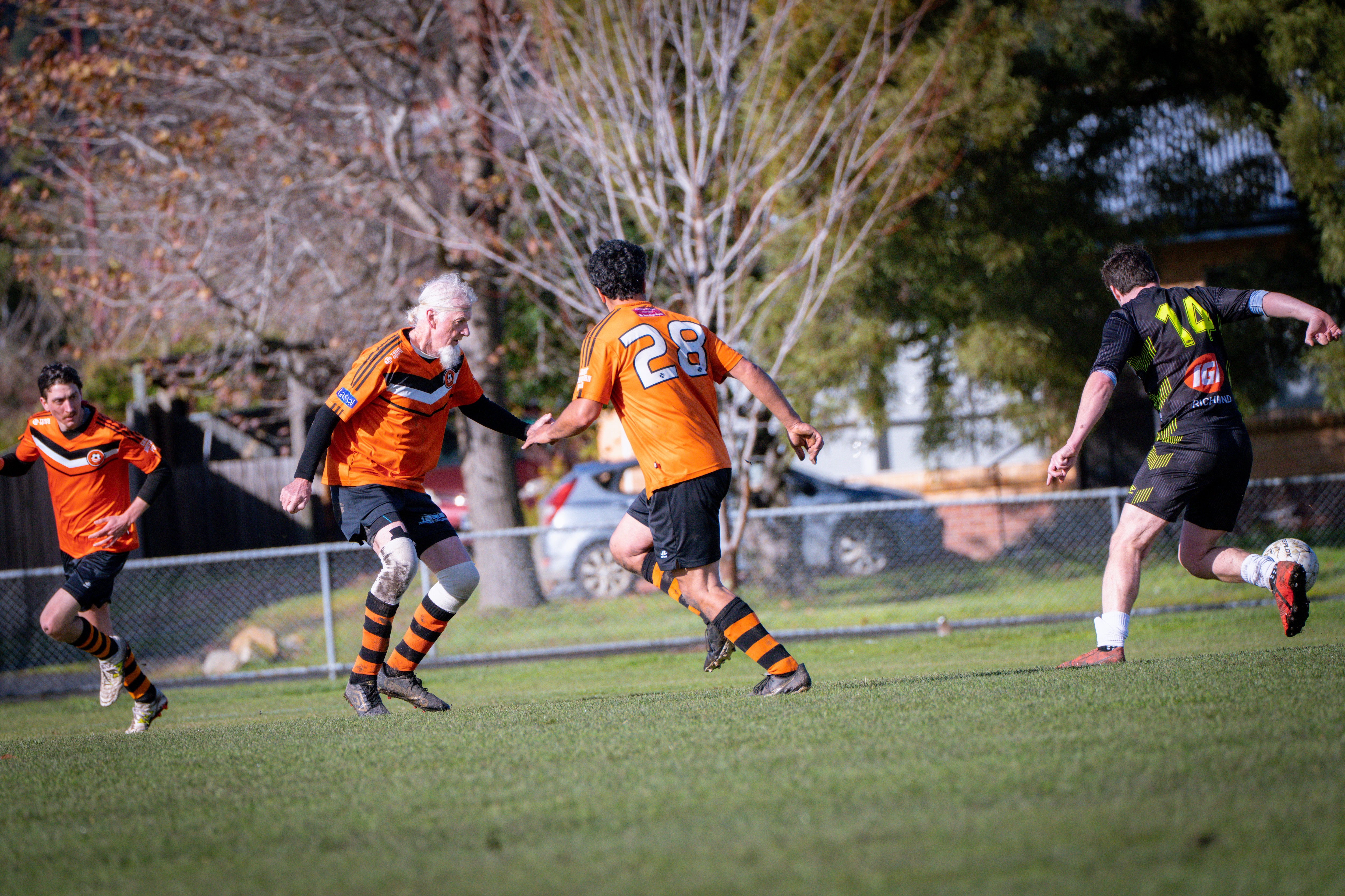 A man with white hair and a long white beard wearing orange soccer colours on a soccer field.
