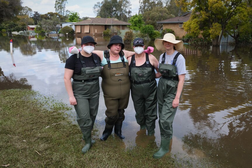 Four women wear gumboots, gaiters, N95 masks and hats as they stand in floodwater with arms around each other. 