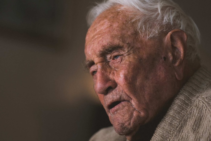 A headshot of a very old man in a tan jumper against a black background.