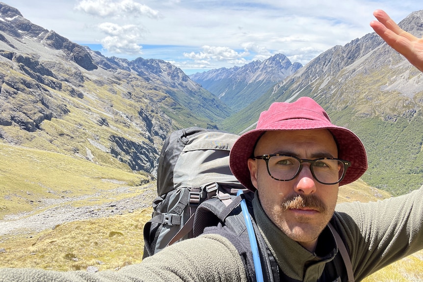 A man with glasses wearing a red hat taking a selfie with mountain ranges in the background.