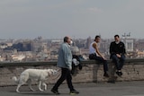 A man walks a dog as a couple sits on a wall at the Gianicolo terrace overlooking Rome.