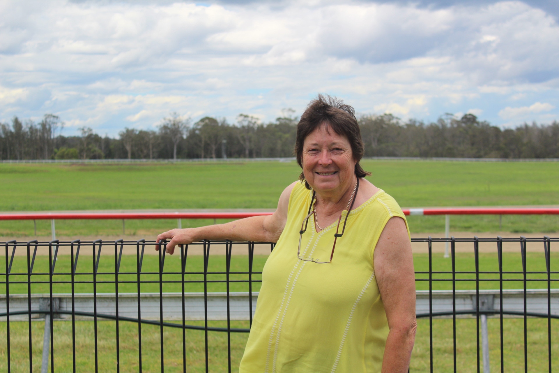 A woman at the fence at a racecourse, wearing a yellow shirt, on a cloudy day.