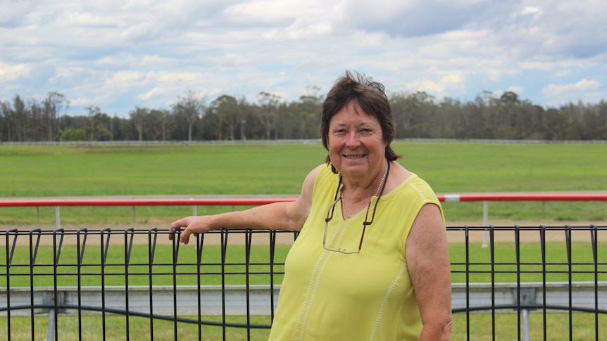 A woman at the fence at a racecourse, wearing a yellow shirt, on a cloudy day.