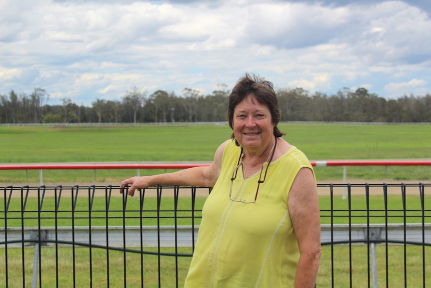 A woman at the fence at a racecourse, wearing a yellow shirt, on a cloudy day.
