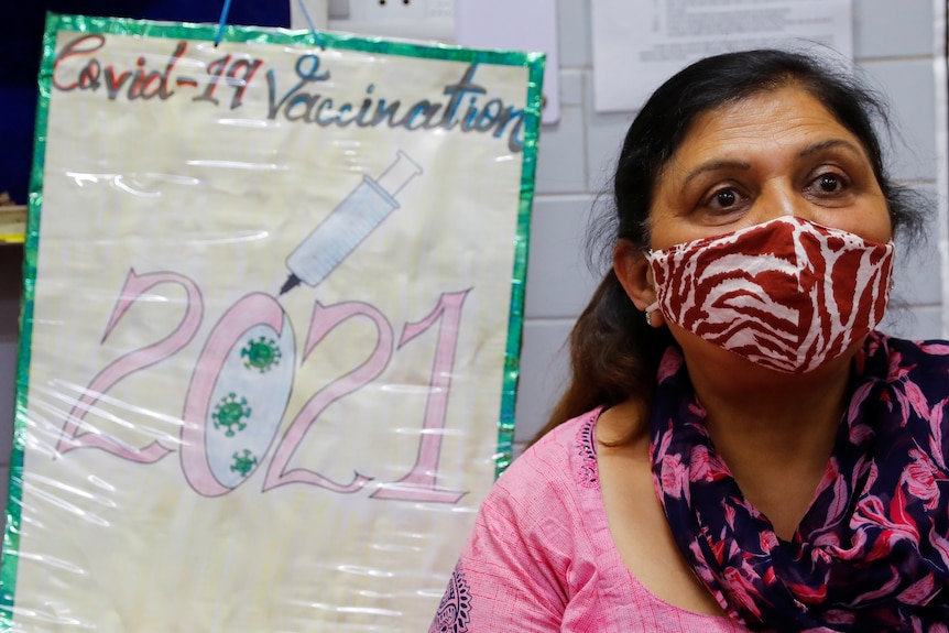 A woman wearing a mask standing in front of a hand drawn poster that says Covid-19 vaccination 2021.