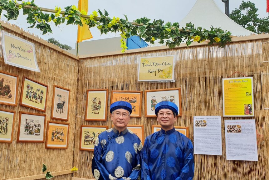 Two men in blue traditional clothes in front of some paintings.
