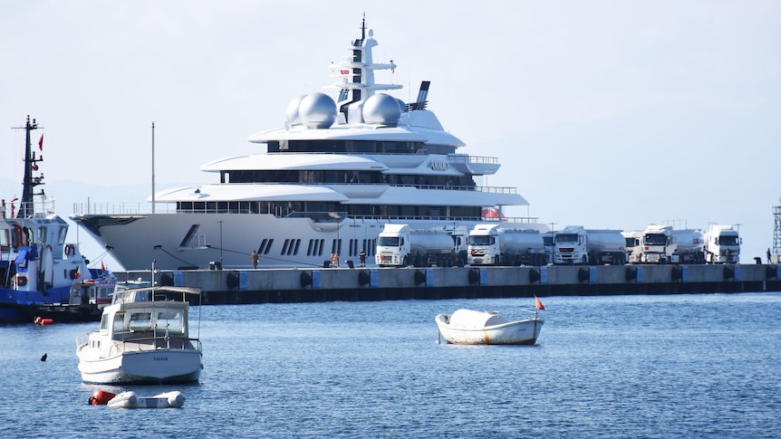 A large superyacht parked next to a wharf, on which sit numerous large trucks. Smaller boats in foreground.