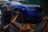 A man attaches an orange ratchet strap to a blue luxury car. 