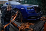 A man attaches an orange ratchet strap to a blue luxury car. 