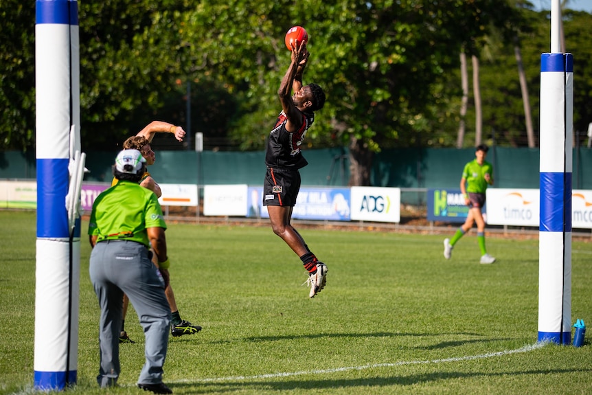 A man playing Australian Rules football leaps to mark a ball in front of the goal posts
