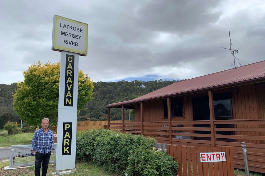 A man wearing a blue flannel shirt stands next to a caravan park sign in north-western Tasmania.