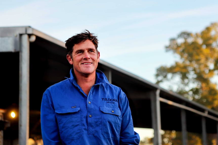 Man wearing blue collared shirt smiles standing in front of woolshed in early morning light with tree visible in background
