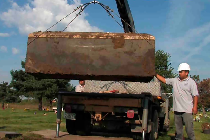 A worker in a hardhat next to a crane lifting a coffin or box.