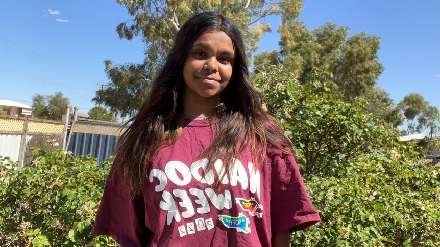 A young woman with brown skin and brown hair smiles wearing a red shirt that says NAIDOC week