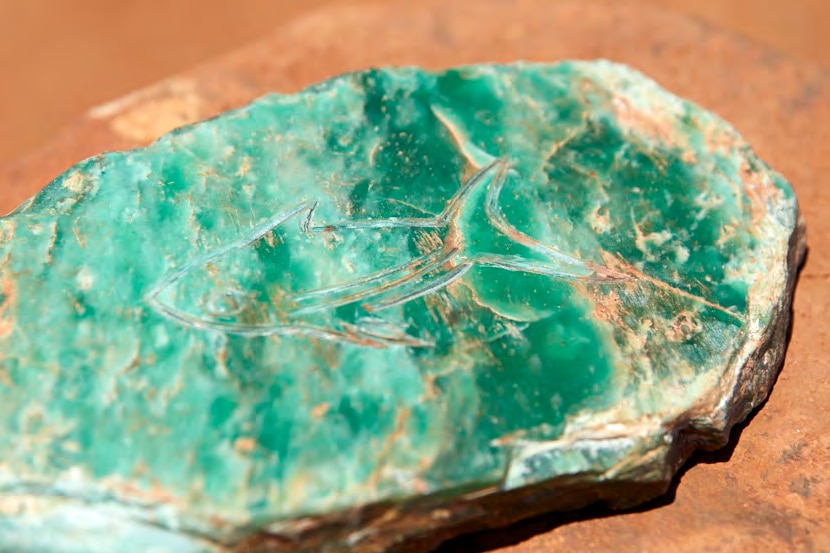 A weathered jade coloured flat stone with simple outline engraving of shark lies on red desert ground on sunny day.