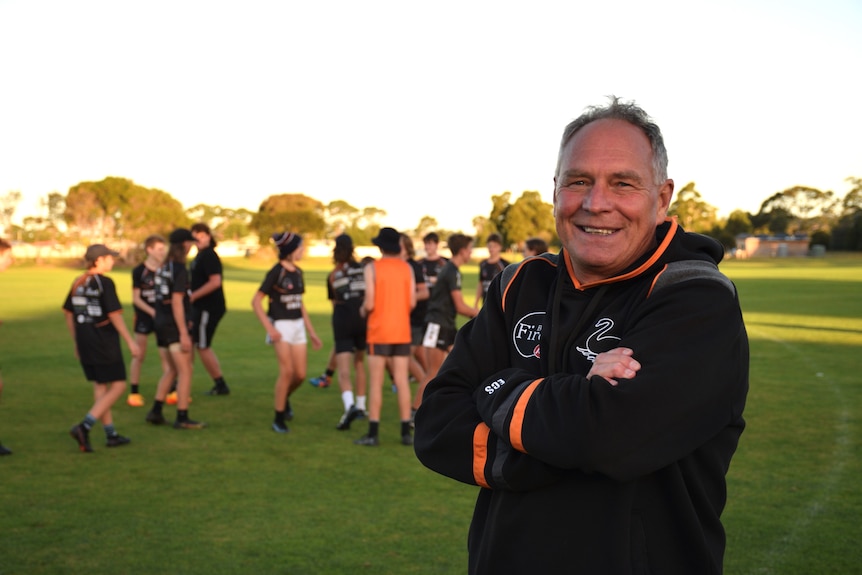 Stephen smiles at the camera while football players gather on the field behind him.