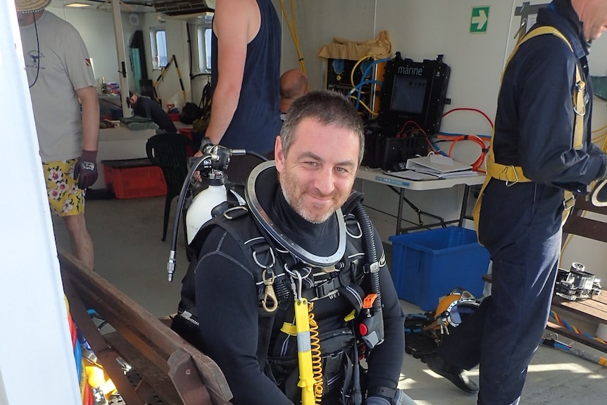 Decked out in diving gear, a dark-haired man with a grey beard sits on boat.