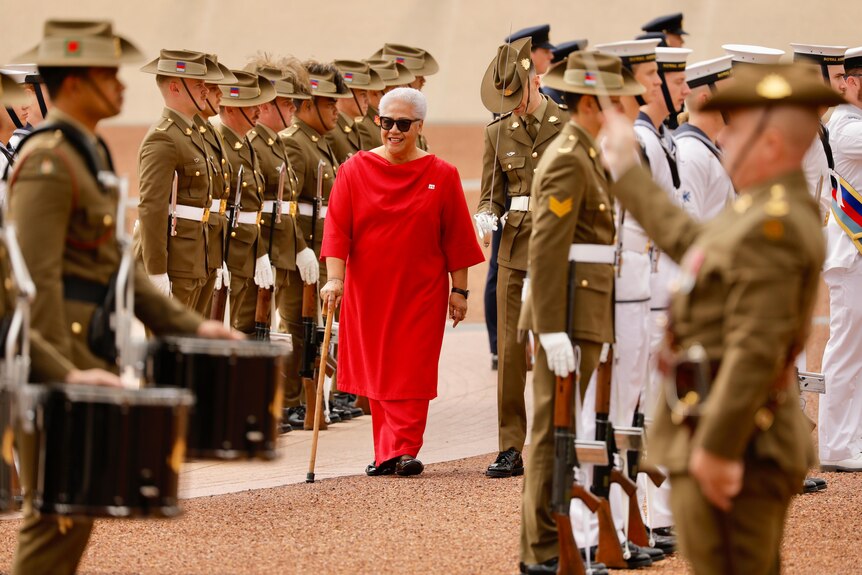 A woman in a red outfit moves with a walking stick through ranks of soldiers at attention in khaki dress uniform.