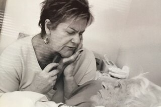 An older woman laying down holds her hand to the face of her daughter, who's looking down on her.