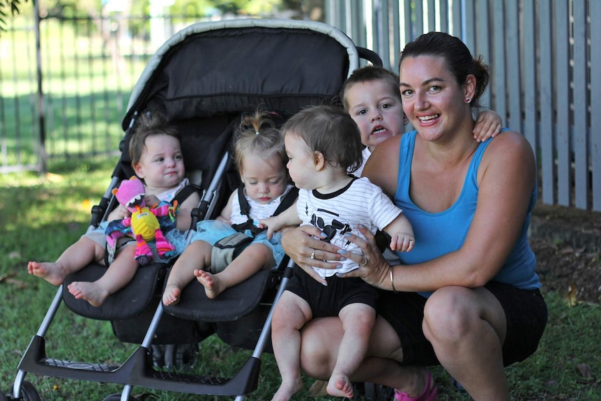 A woman in sports gear squats on the ground in front of a fence with her four children, three of which are triplets.