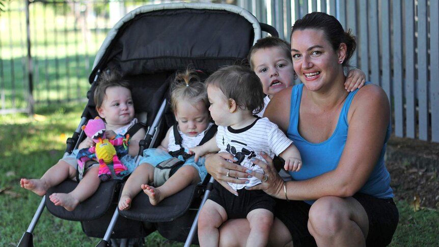 A woman in sports gear squats on the ground in front of a fence with her four children, three of which are triplets.