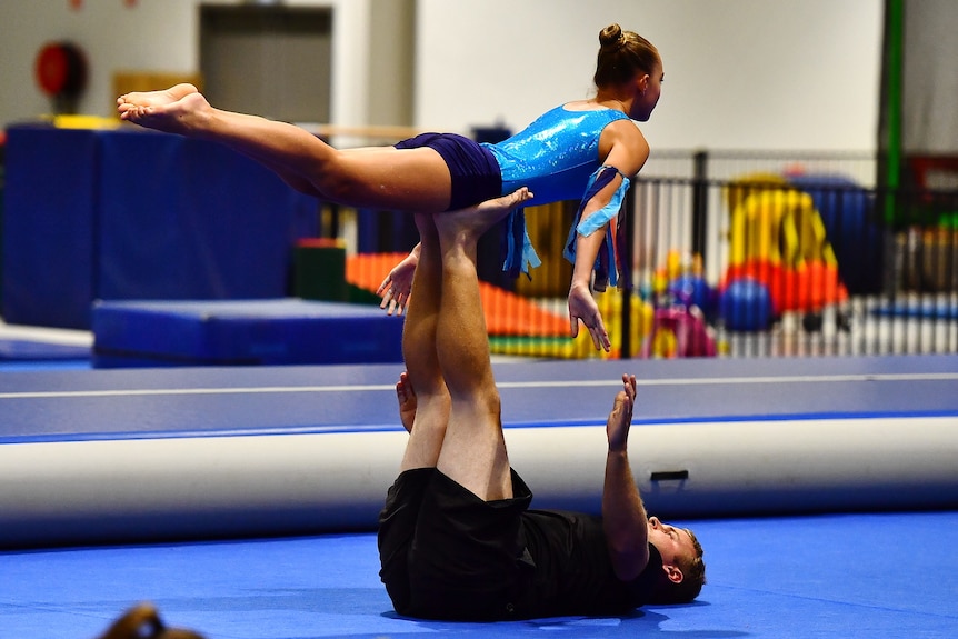 A young woman wearing a leotard balances on a young man's legs in a gymnasium.