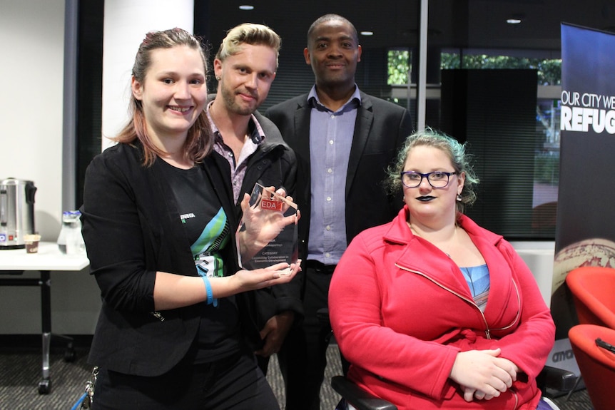 Four people smile at the camera as the young woman at the front holds up a glass trophy.