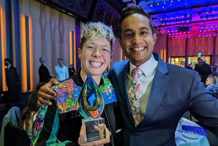 Mon at an awards night, holding a trophy and smiling. They are standing with Jeremy Fernandez.