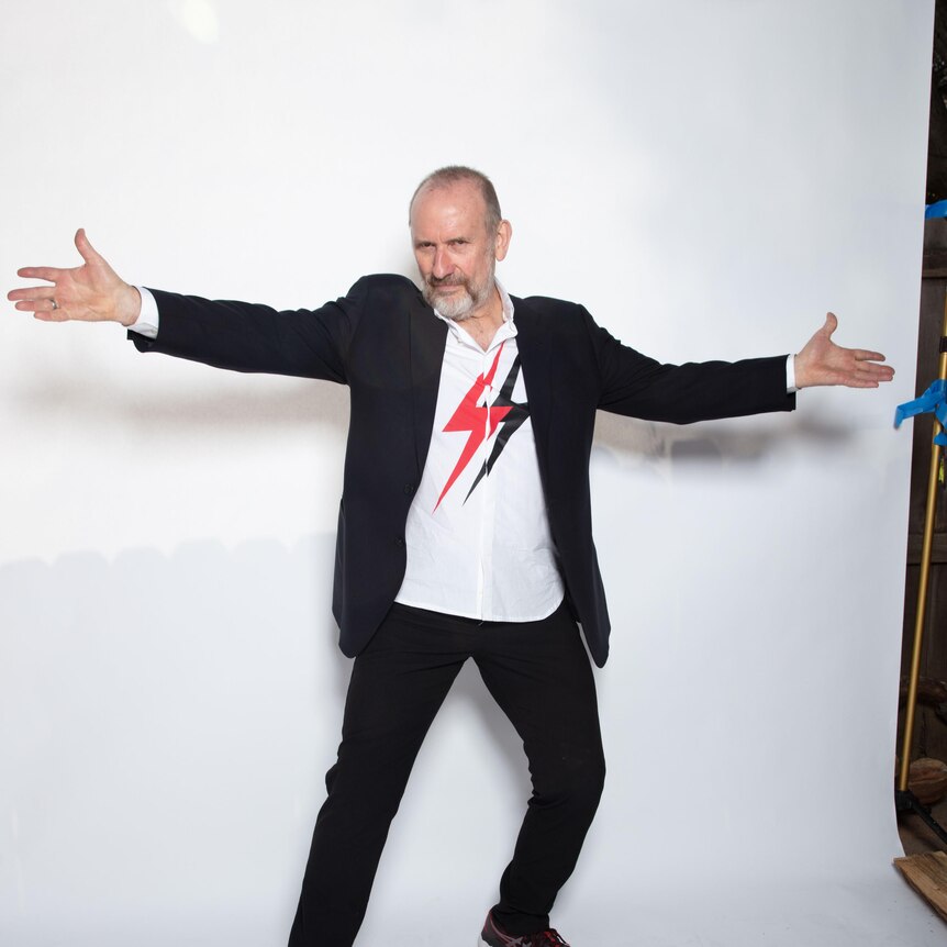 Singer Colin Hay standing with his arms outstretched in a welcoming gesture