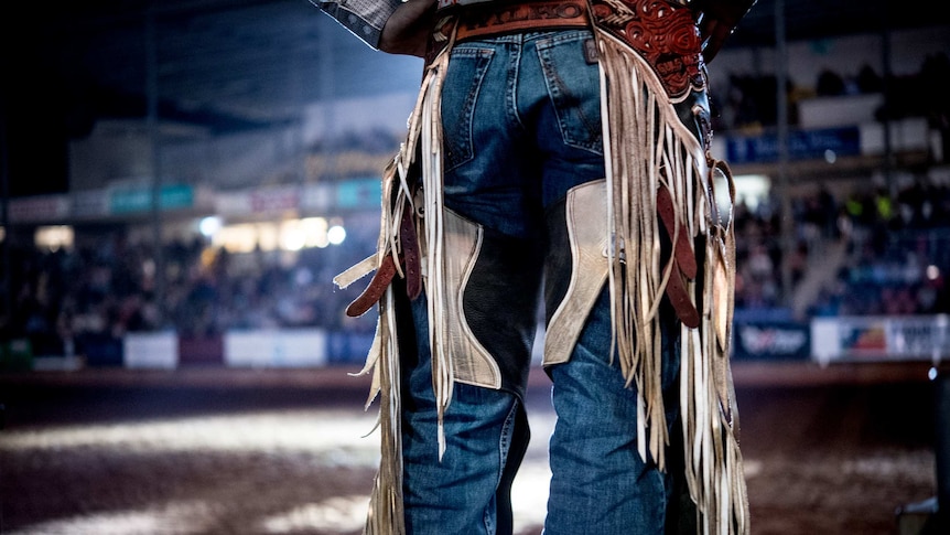 A spotlight shines into the rodeo arena at night with a bull fighter's legs in the foreground.