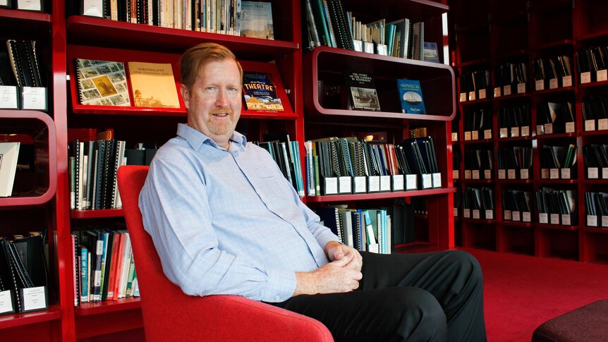 A man with red hair sits in a red chair surrounded by red bookshelves filled with books and files.
