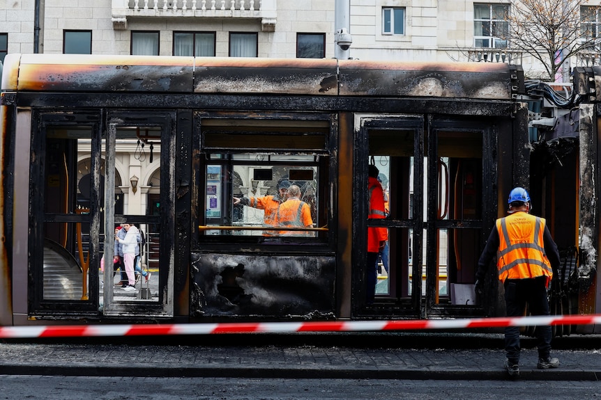 A burned metal hull of a tram with people wearing orange around it and a police tape in front
