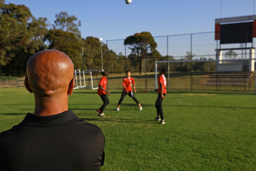 Man looks on as three junior soccer players kick a soccer ball on the field.