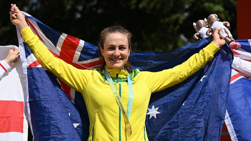 An Australian female road cyclists stands with the national flag behind her while wearing her Comm Games gold medal.